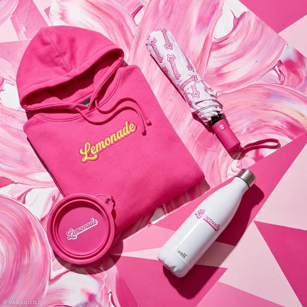 קיט לייף-סטייל ממותג של Lemonade הכולל קפוצ'ון ורוד, מטרייה תואמת, בקבוק שתייה לבן וקערת כלבים ורודה על רקע גרפי ורוד.