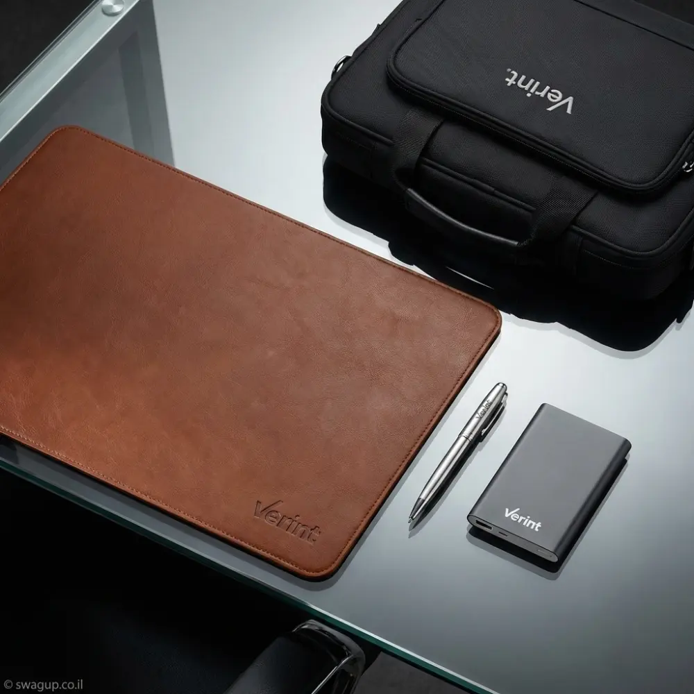 קיט מיתוג יוקרתי של חברת Verint הכולל משטח עבודה מעור חום, תיק מחשב שחור, סוללת גיבוי ועט מתכת על שולחן משרדי שקוף.