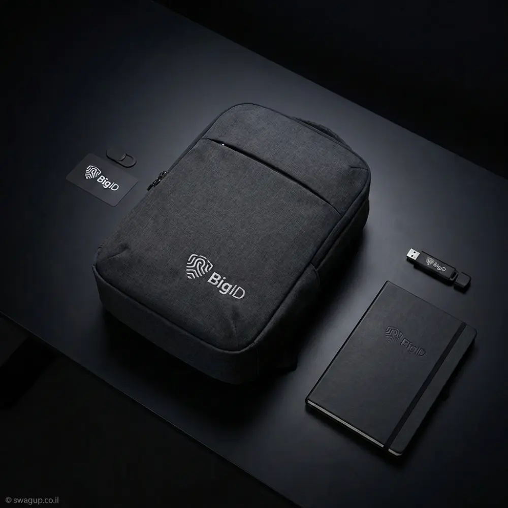 קיט מיתוג טכנולוגי של חברת BigID הכולל תיק גב שחור, מחברת כריכה קשה, כיסוי מצלמה וזיכרון נייד על שולחן עבודה כהה.