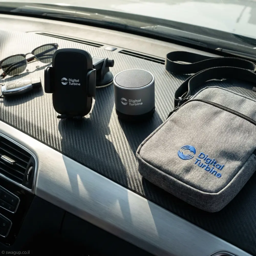 קיט מיתוג של חברת Digital Turbine הכולל תיק צד אפור, רמקול בלוטות' שחור ומעמד לטלפון על רקע לבן נקי.